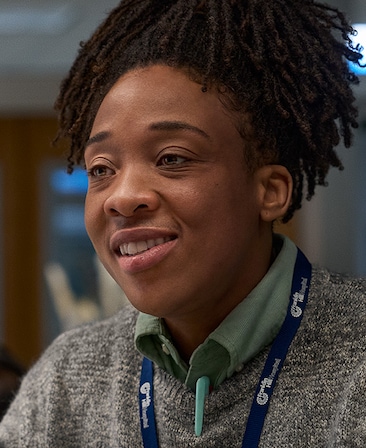Headshot of Esco Jouley playing Sonya, wearing a grey sweater with a blue lanyard on.