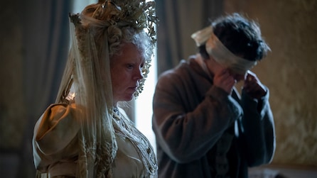 Olivia Coleman looking down and standing next to a man that is blindfolded in room with curtains