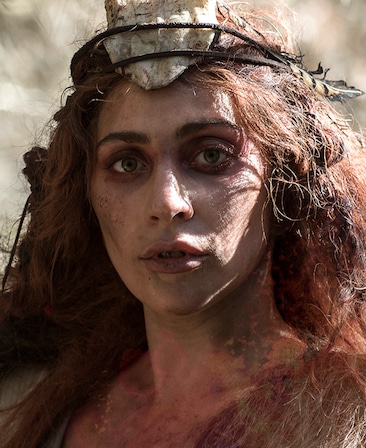Lady Gaga with long red hair and dirt caked on face and neck and wearing leaf and twig crown with rock centerpiece