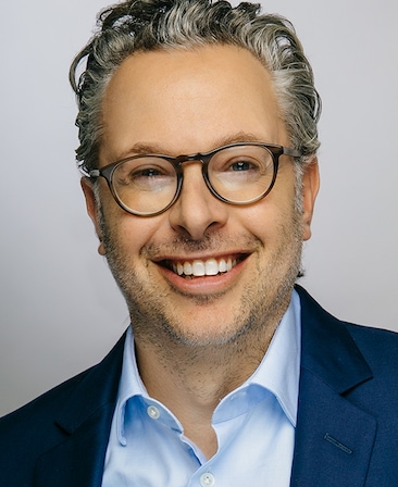 Headshot of Aaron Hart wearing a light blue button up and dark blue blazer, paired with brown rounded glasses.