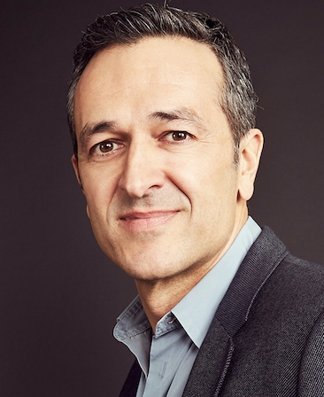 Headshot of Hernan Lopez wearing a blue button up, grey suit, with a grey background behind him.