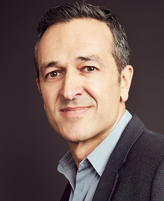 Headshot of Hernan Lopez wearing a blue button up, grey suit, with a grey background behind him.