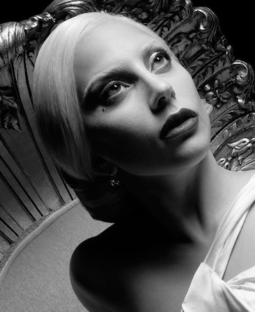 Black and white headshot of Lady Gaga with slicked back white hair and white eyebrows, looking up in white shoulder dress