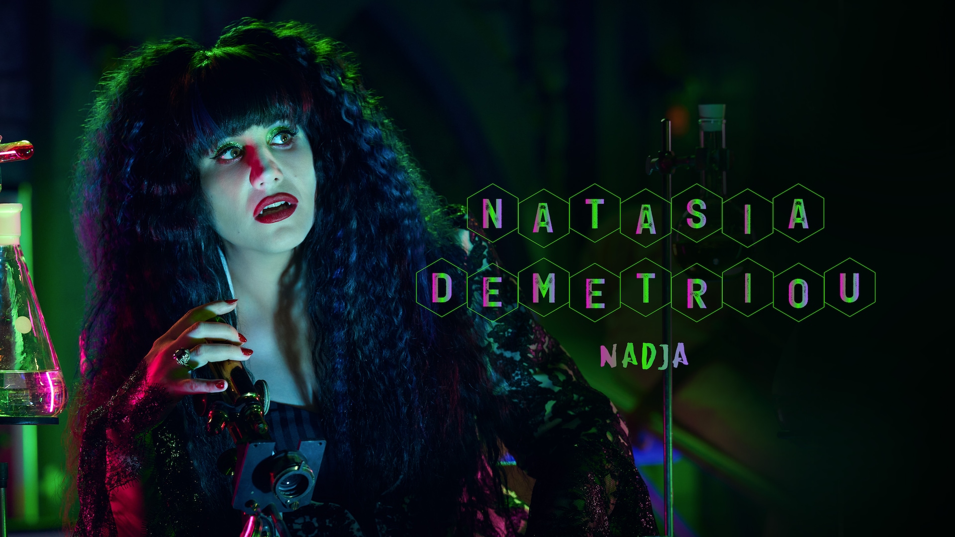 Natasia Demetriou as Nadja headshot wearing red lipstick with her hair crimped holding a microscope in a laboratory.