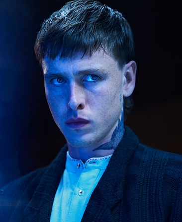 Harris Dickinson headshot wearing a white shirt under a black jacket with a tattoo on his neck