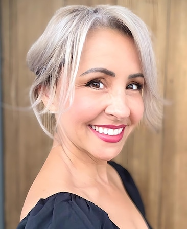 Headshot of Nikki Boyer wearing a black ruffled top, hair tucked into a bun, standing in front of wood paneling.
