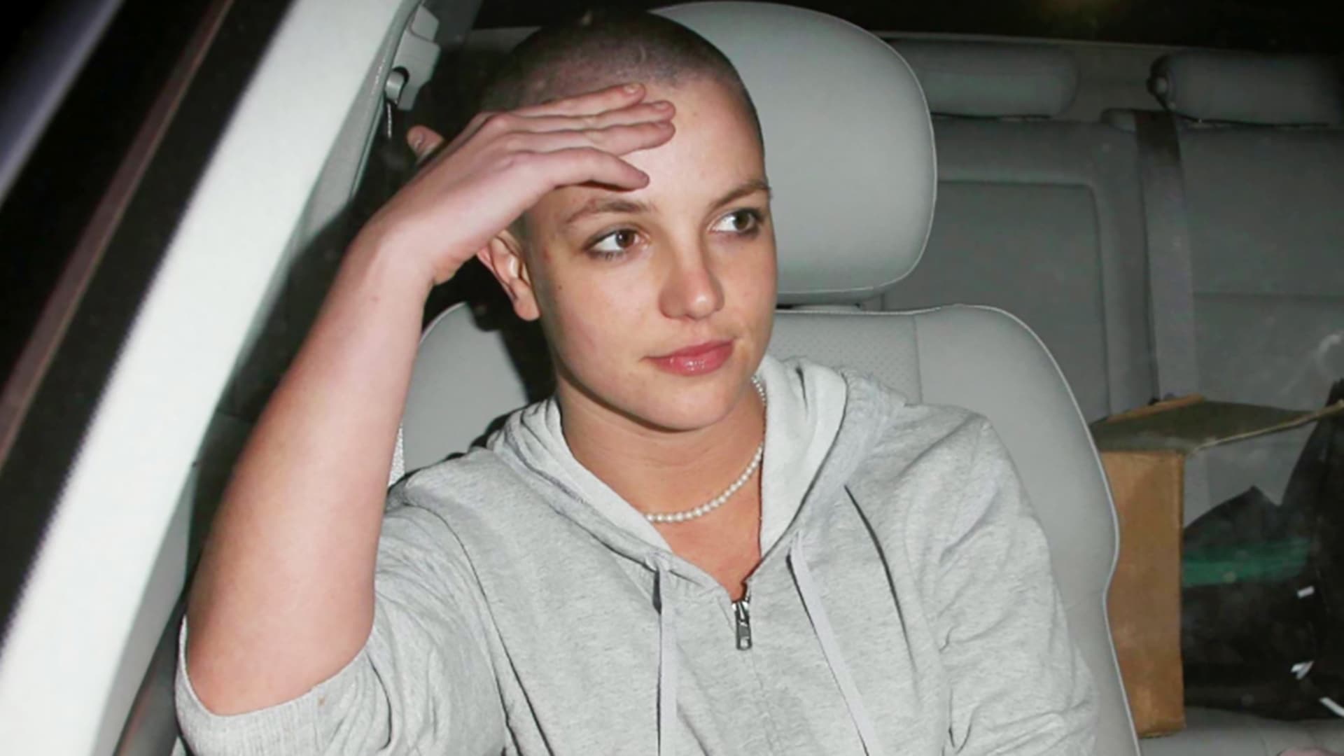 Britney Spears sitting in the passenger seat of a car wearing a grey hoodie. 