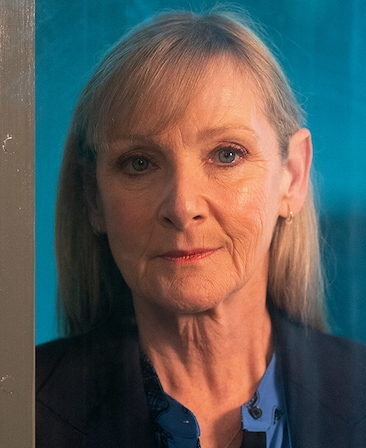 Lesley Sharp headshot wearing a blue shirt and navy jacket in front of a blue background