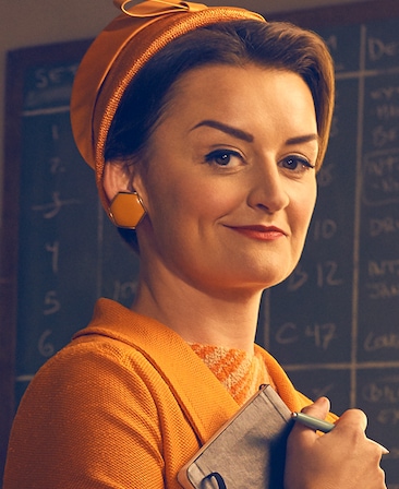 Alison Wright as Pauline Jameson in orange hat earrings and blazer holding notebook and pen by blackboard in Feud FX show