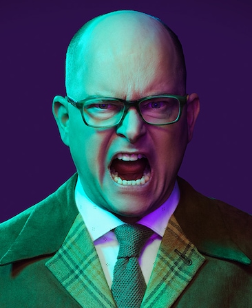 Mark Proksch headshot opening his mouth wide, wearing glasses and a plaid collar suit jacket.