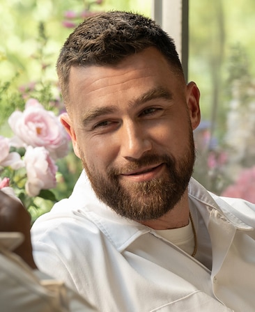 Travis Kelce as Ed Laclan in FX's Grotesquerie, wearing a white button up, sitting on a couch with a flowers behind him.