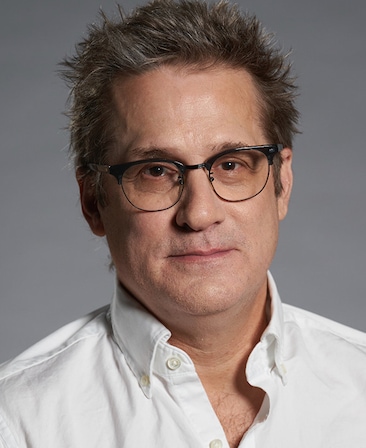 Headshot of executive producer Paul Simms wearing a white button up, with a grey background.