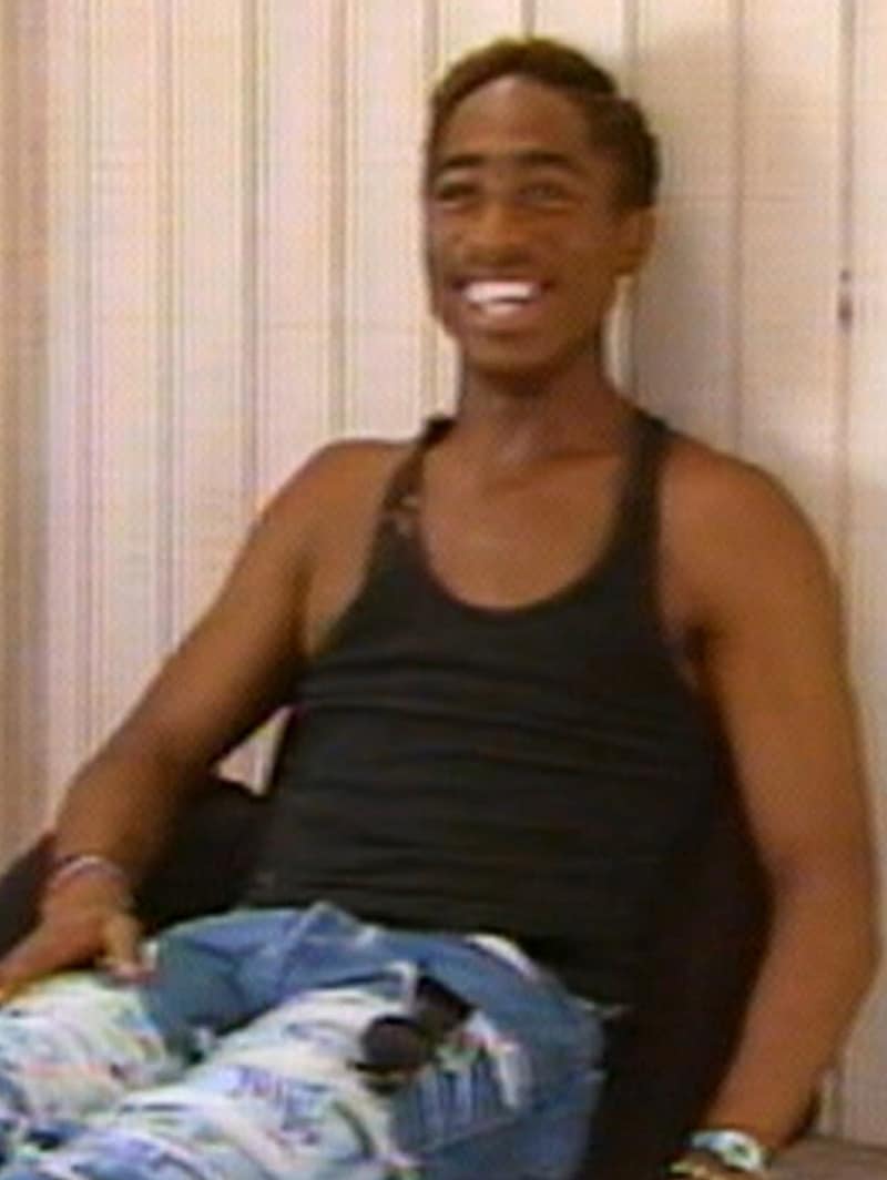 Tupac Shakur wearing a black tank top and blue jeans leaning against a white wall