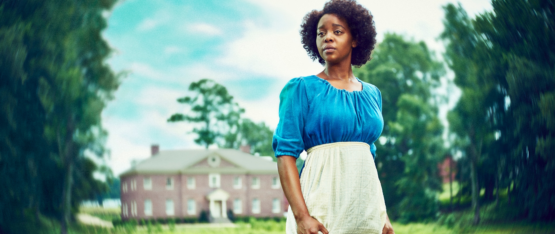 Mallori Johnson as Dana for FX's Kindred wearing a blue dress and white apron with a brick manor behind her. 