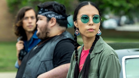 Girl wearing a green jacket and blue tinted sunglasses with a circle frame, leaning against a car outside.