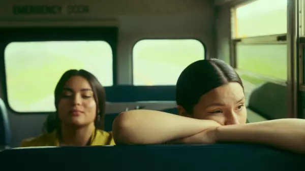 Elora and Willie sitting on a school bus, as Willie leans on the seat in front