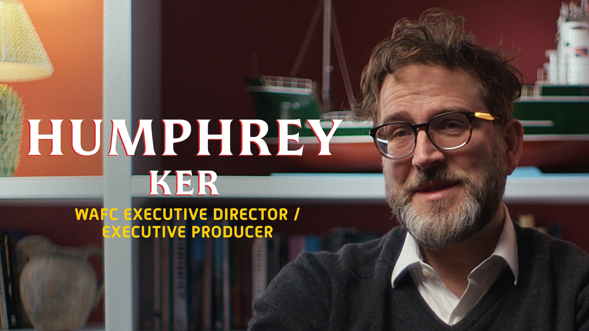Humphrey Ker headshot wearing a white button up under a grey sweater, wearing tortoise printed glasses and his name in white.