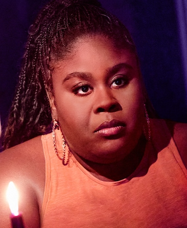 Raven Goodwin as Merritt Tryon in FX's Grotesquerie in an orange shirt, gold hoops, holding a burgundy candle in a dim room.