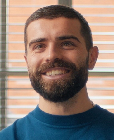 Elliot Lee headshot wearing a blue t-shirt, standing in front of a window