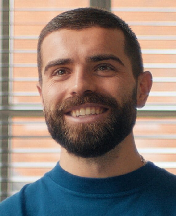 Elliot Lee headshot wearing a blue t-shirt, standing in front of a window