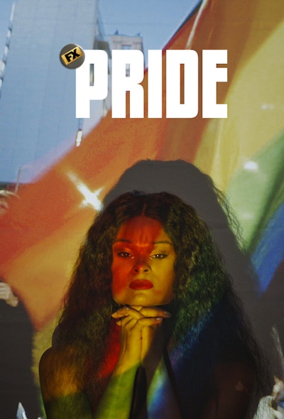 Woman in front of a projector displaying a PRIDE parade with masked participants, and the rainbow flag colors on her face