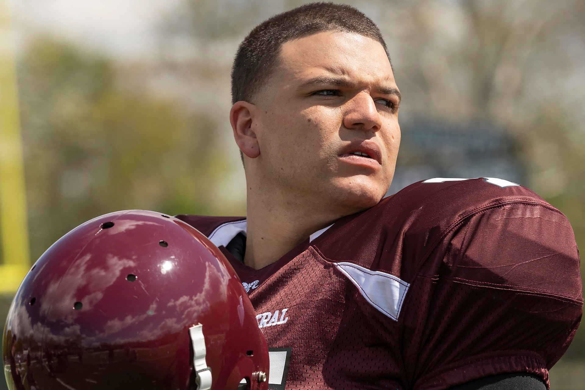 Hernandez wearing a burgundy football uniform and holding a helmet looks to the side of the field