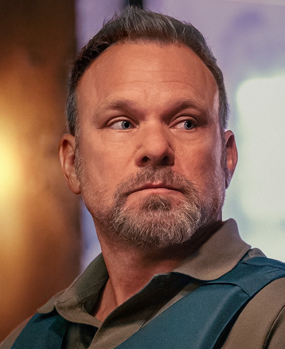 Norbert Leo Butz headshot wearing a brown shirt and blue padded vest