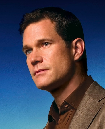 Dylan Walsh headshot wearing a brown suit jacket and shirt against a blue background