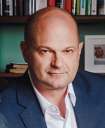 Headshot of Brad Simpson wearing a blue suit in front of a bookshelf.