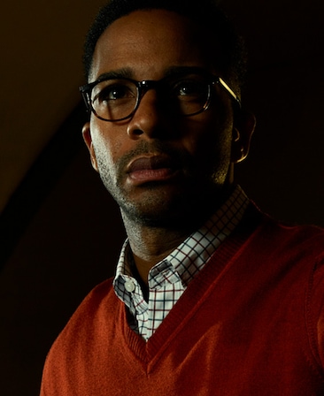André Holland headshot in checkered white button down and red v neck sweater and glasses in dark room with half face shadowed