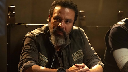Bishop wearing a leather vest that reads "Mayans" and clasping his hands together.