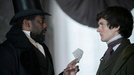 Ashley Thomas in black suit and top hat handing Fionn Whitehead a written piece of paper