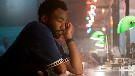 Donald Glover as Earn at table on his elbows with hand on face, surrounded by rainbow lights and green lamp in FX's Atlanta