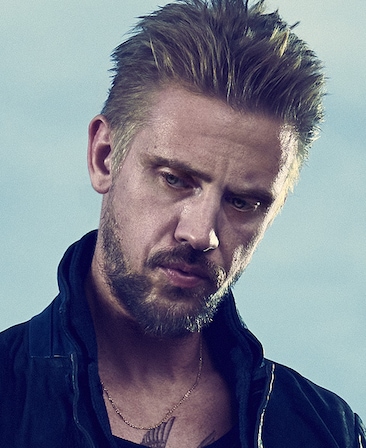 Boyd Holbrook headshot wearing a black shirt with a gold necklace