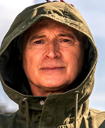 Robert Carlyle headshot wearing a green jacket with a hood over his head
