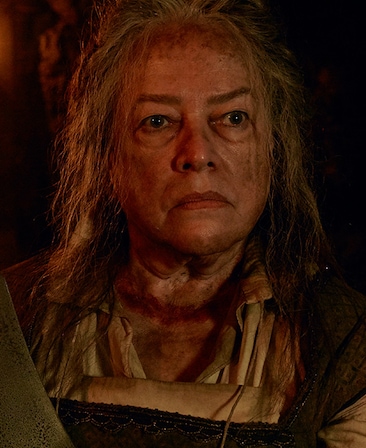 Kathy Bates with disheveled hair, dirt and grime on face, in dirtied white peasant shirt and holding machete knife