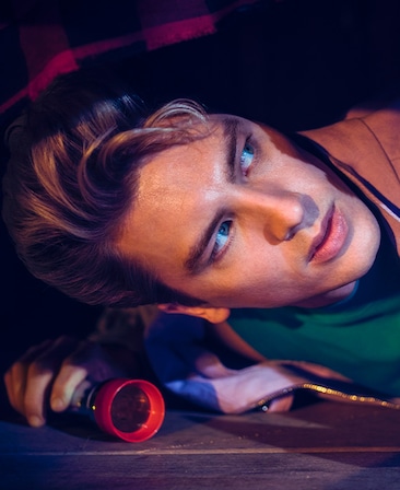 Cody Fern headshot lying down on wooden floor and looking up while holding flashlight from American Horror Story 1984
