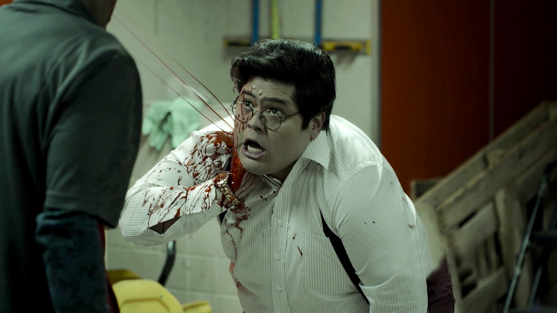 Guillermo covering his neck spurting neck wound in pain, wearing a white button up in the backroom of a store.