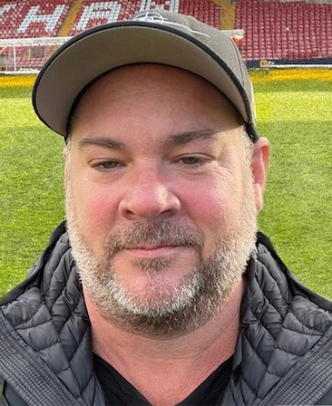 Josh Drisko headshot wearing a black puffer jacket and baseball cap, standing outside on the soccer field