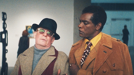 Truman Capote wearing a hat and sunglasses while standing next to a man with an orange coat.