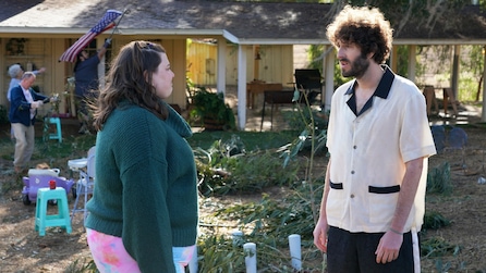 Dave Burd outside in front of a house wearing a white and black blowing shirt talking to a girl in a green sweater