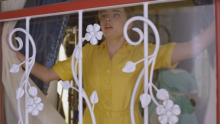 Scared woman in vintage yellow button down dress pulls back window covers in American Horror Stories Installment 2