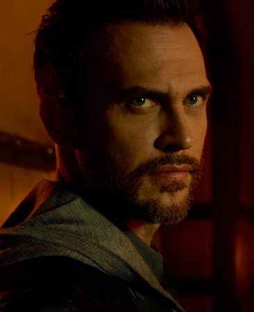 Cheyenne Jackson headshot with trimmed beard wearing grey hoodie and looking over his right shoulder in dark room