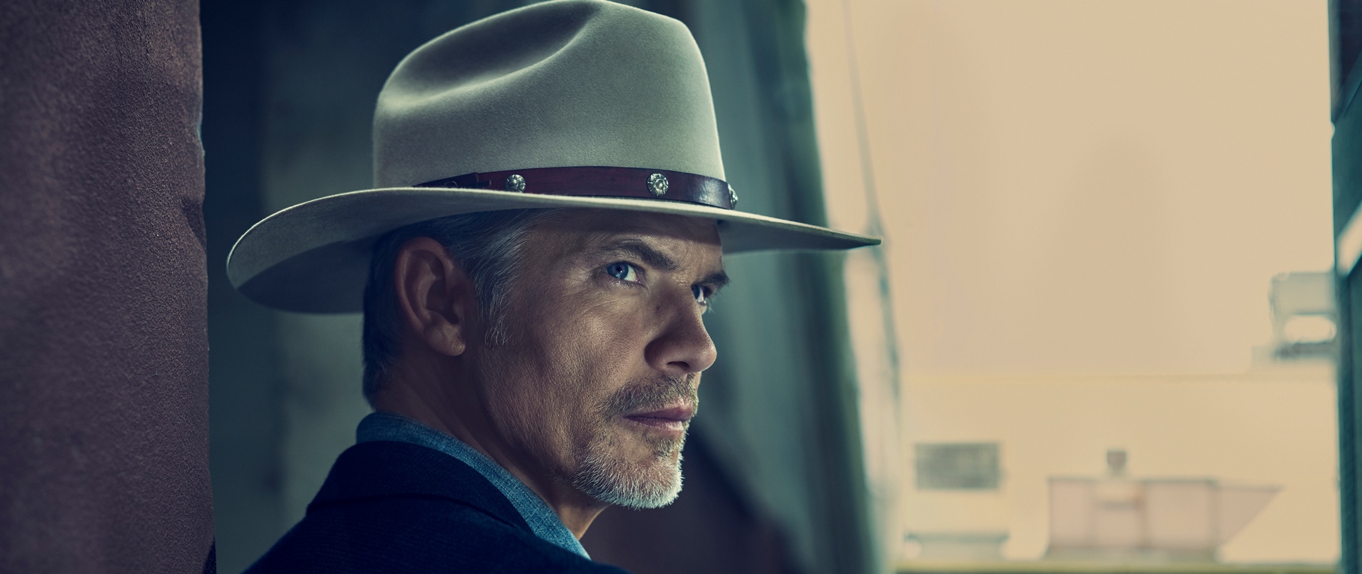 Timothy Olyphant as Raylan Givens wearing a tan hat in a blue colored shirt looking off to his side.