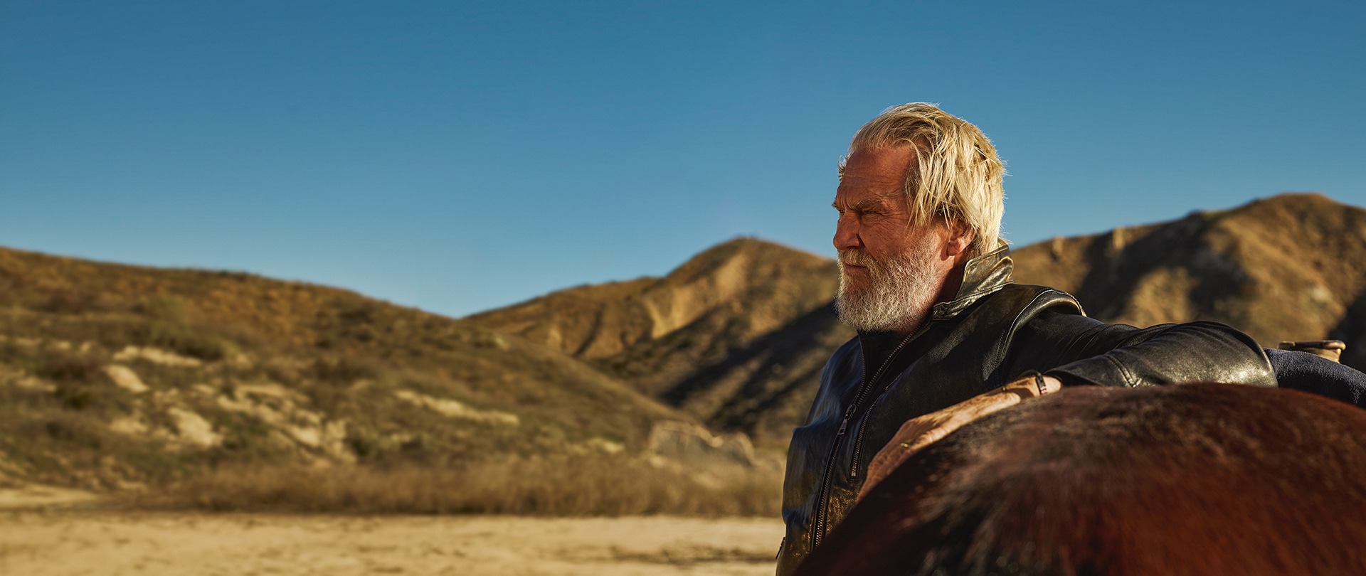 Jeff Bridges as Dan Chase in a leather jacket with is arm resting on a horse amidst the desert.