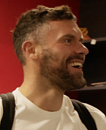 Ben Foster headshot wearing a white shirt and black backpack over his shoulders, standing against a red background