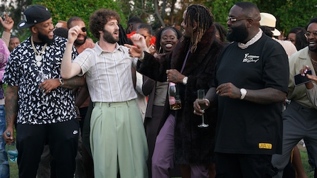 Dave Burd wearing green trousers and a gray top standing next to a group of people