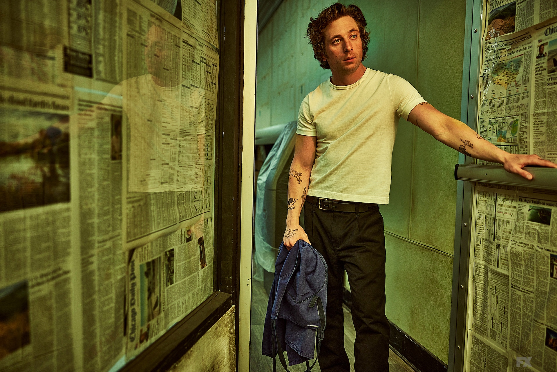 Jeremy Allen White as Carmy wearing a white shirt and black pants, leaning his hand on a door covered in newspaper
