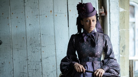 Estella wearing a purple dress and hat standing outside by white wooden wall and holding black stick