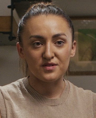 Phoebe Davies headshot, wearing a brown sweater with her hair tied back, sitting in front of a frame on the wall.